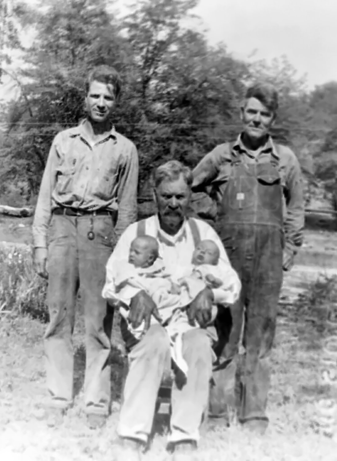 Four generations in a yard: an old man with a moustache sits in a chair holding two newborn twins wrapped in white; a young man in a work shirt stands at the left and an older man in overalls stands at the right, trees behind them.