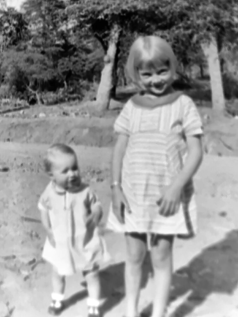 Two children on a dirt yard in sunlight: a laughing girl of about six in a striped dress and a toddler in a white smock, trees behind.