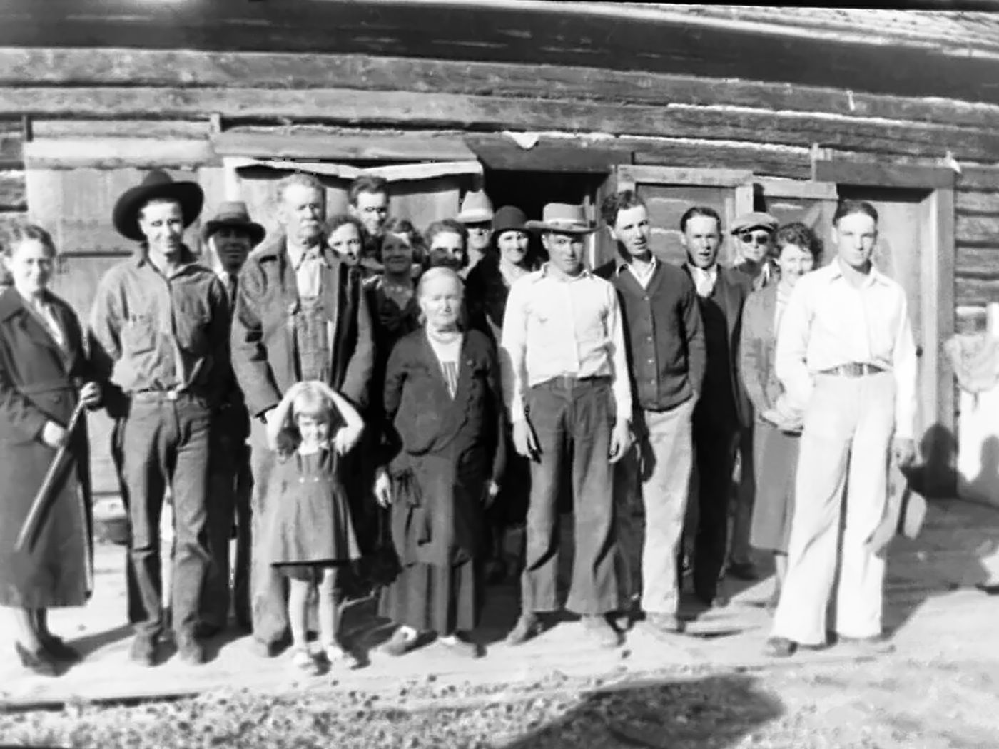 About twenty people, men in hats and work clothes and women in dresses, stand in a row in front of a long log building; a small girl in a pinafore stands at the front with her hands on her head.
