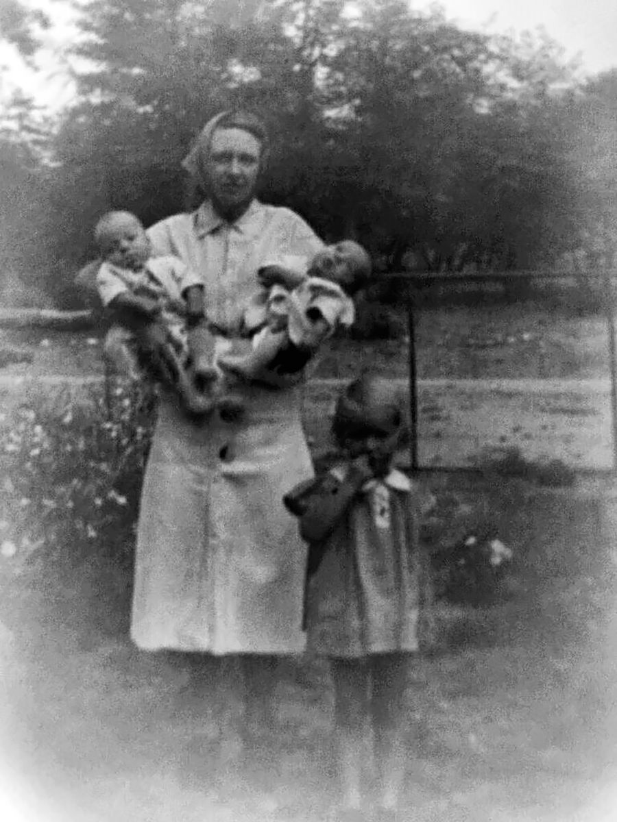 A woman in a headscarf and a light dress holds two infants, one on each arm, in a garden; a small girl in a dark dress stands at her side looking up.