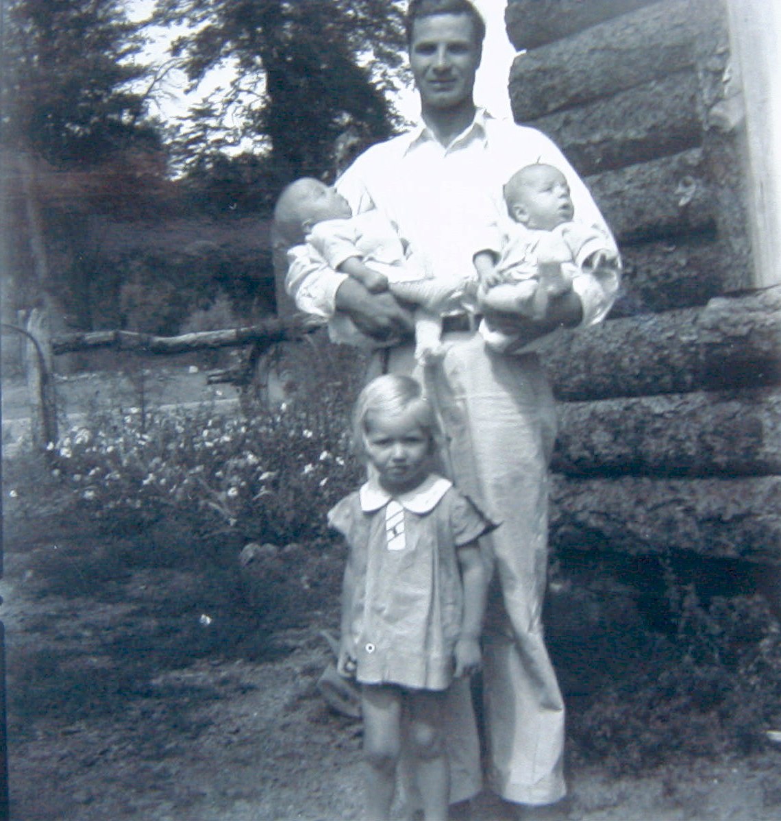 Floyd Waldrip holding infant twins with young daughter Janice outside log cabin